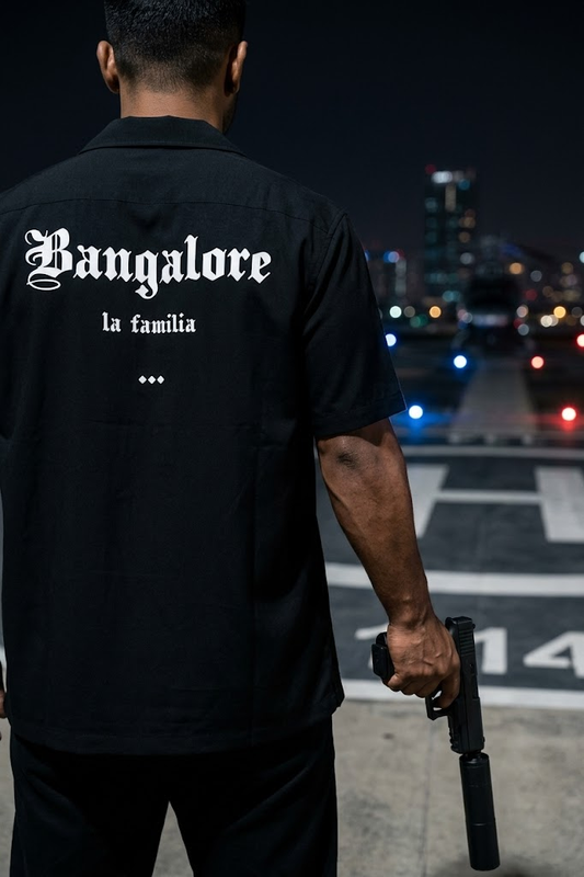 Person wearing a black shirt with 'Bangalore la familia' text, holding a gun on a city street at night.